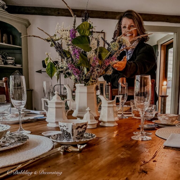 Ann, vintage home decor blogger at Dabbling & Decorating next to a summer into fall table setting with family heirlooms at her Maine home before heading to Vermont for the winter.
