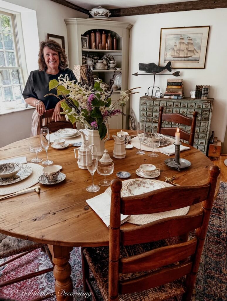 Ann, vintage home decor blogger at Dabbling & Decorating next to a summer into fall table setting with family heirlooms at her Maine home before heading to Vermont for the winter.