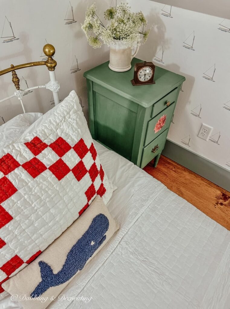 Painted green nightstand on side of antique twin wrought iron bed with red and white sham pillow and whale pillow in attic bedroom.