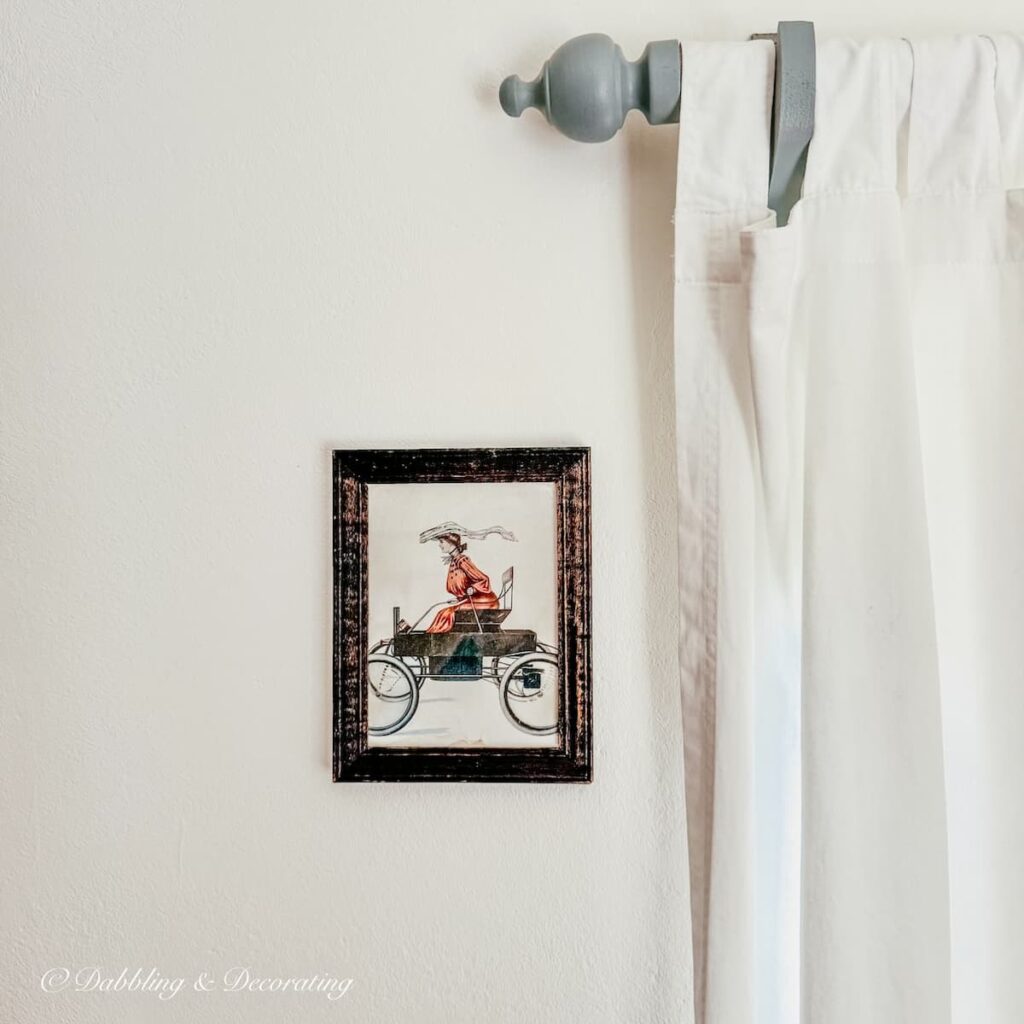 Benjamin Moore's Simply White painted wall with a simple antique black framed picture and white curtains hanging on a blue-gray curtain rod.