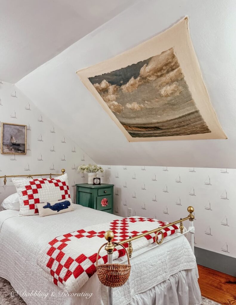 Small attic bedroom with slanted ceilings with a vintage tapestry displayed over an antique wrought iron twin bed with red and white bedding.