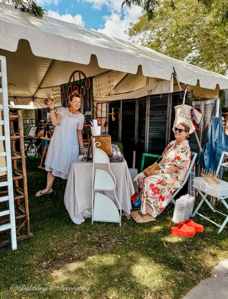 Two women vendors at Van Claus Designs laughing and waving at Vintage Bazaar New England in Mystic, CT from their antique booth display.