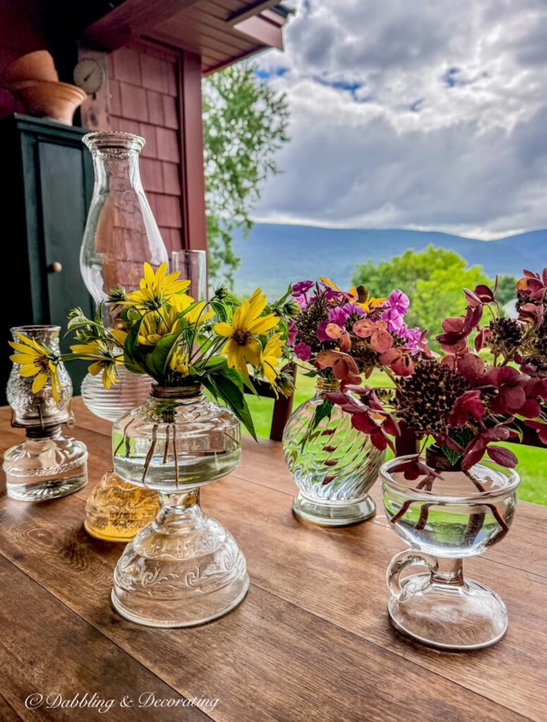 Six antique oil lanterns in different sizes styled on a fall porch table with flowers and chimney's with a mountain view.