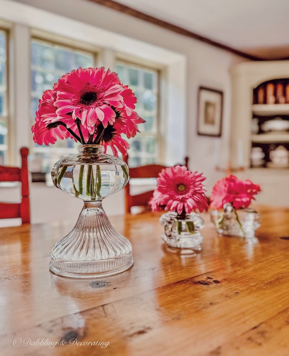 Three antique oil lamps in different sizes and shapes repurposed into flower vases on dining room table with pink daisies.