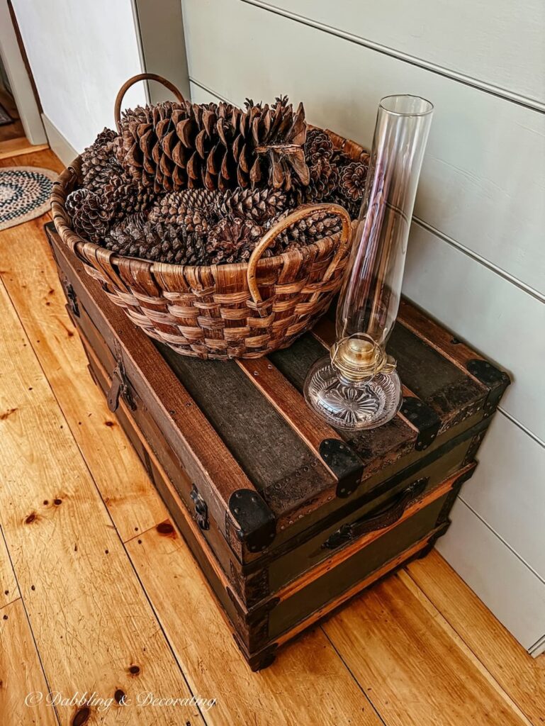Large round basket filled with pinecones resting on top of an an antique trunk and an antique oil lamp next to it.