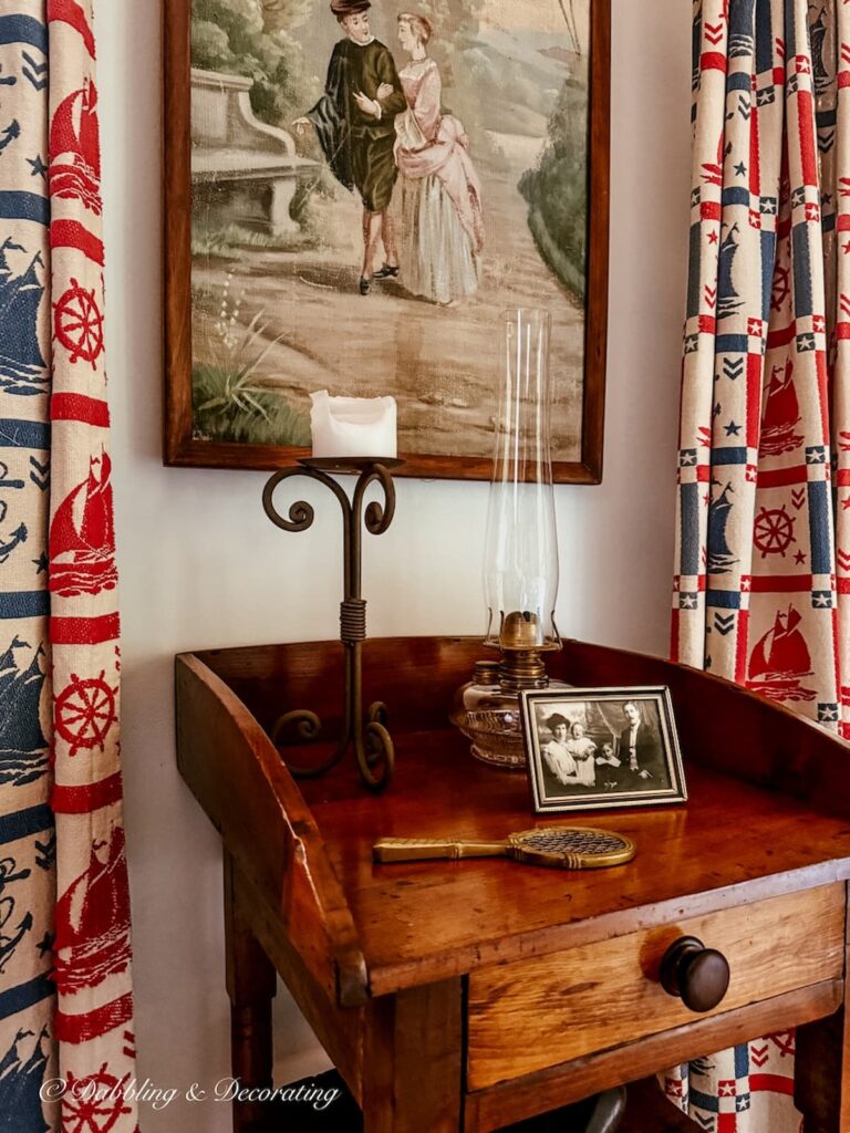 Antique wooden side table styled between windows with an antique oil lamp, candle, picture and wall art.