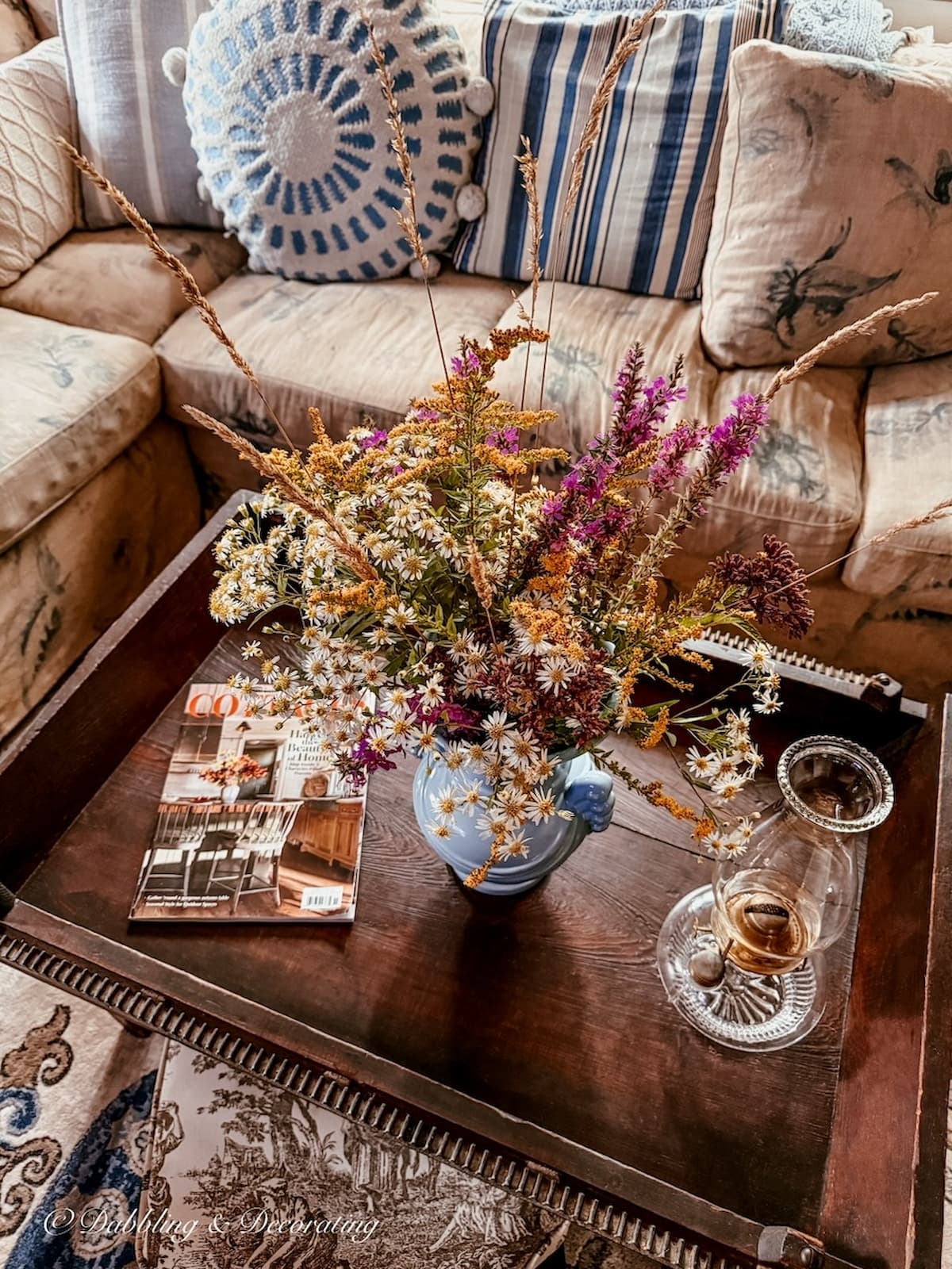 Antique butter churn coffee table styled with a bouquet of wildflowers, magazine and an antique oil lamp in vintage home's living room.