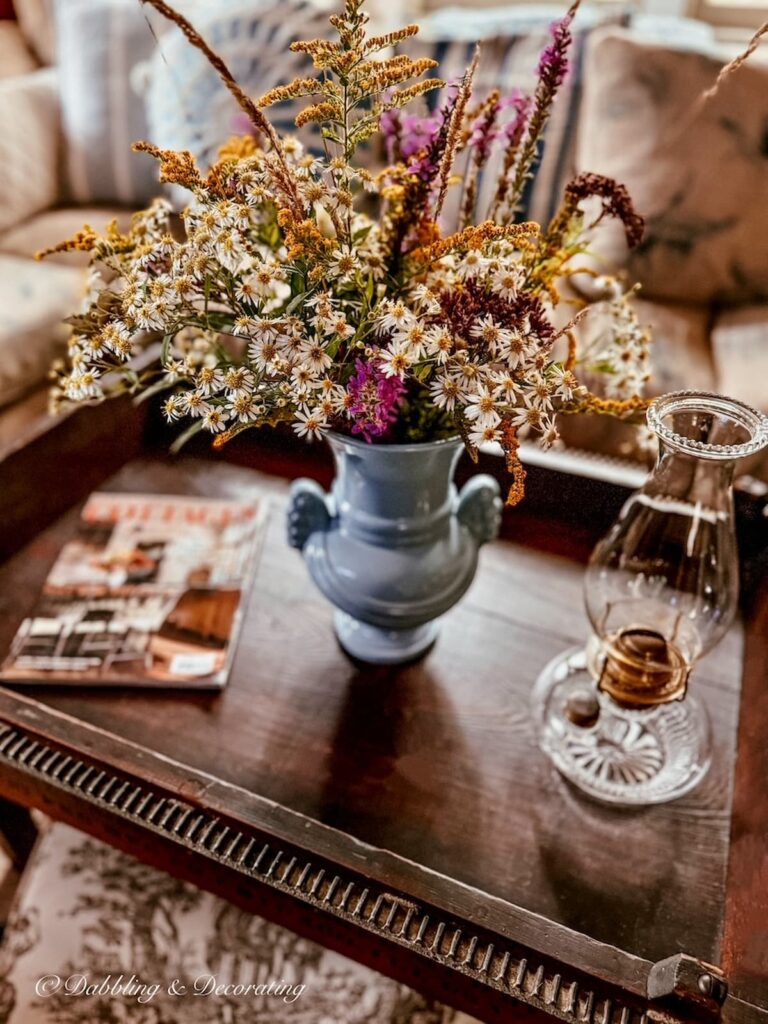Antique oil lamp styled on antique wooden coffee table with wildflowers in blue vase and magazine in vintage home's living room.