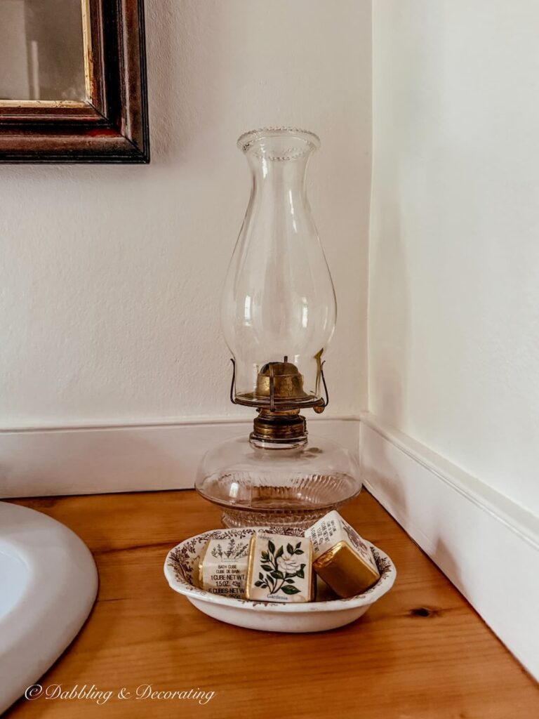 Antique oil lamp on wooden bathroom countertop displayed with a vintage transferware soap dish with wrapped soaps next to sink and vintage wall mirror.