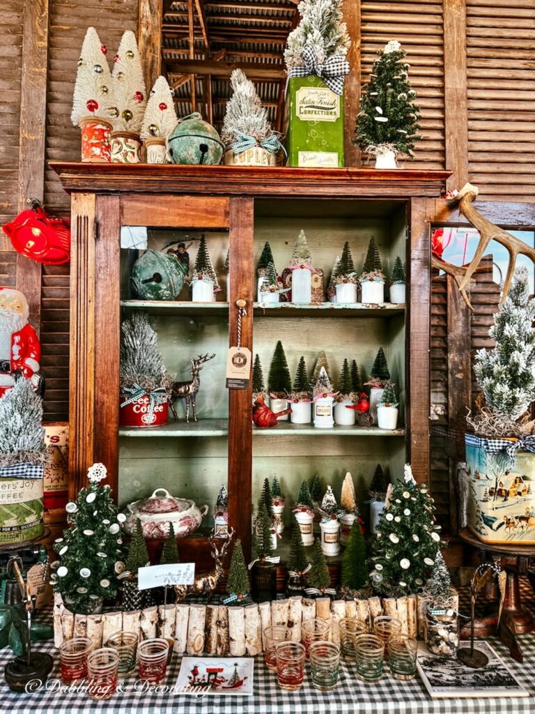 Vintage wooden apothecary cabinet filled with small bottle brush Christmas trees inside a vintage Christmas booth display.