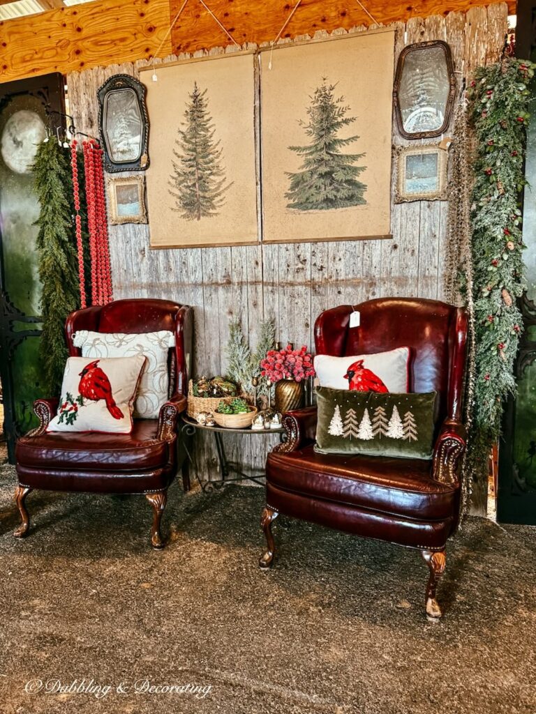 Two red leather chairs in a vintage booth Christmas display with barn wood wall decor, Christmas garlands, vintage mirrors, red cardinal pillows and Christmas tree wall paper drops at the Country Living Fair.