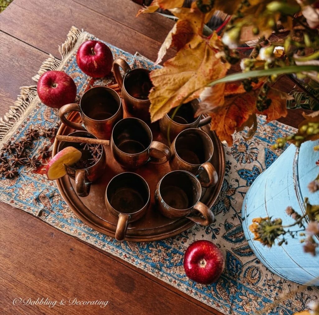 A set of 8 vintage copper mugs on a copper tray styled on an outdoor Vermont porch table with an antique Iranian textile and a foraged fall flower arrangement.