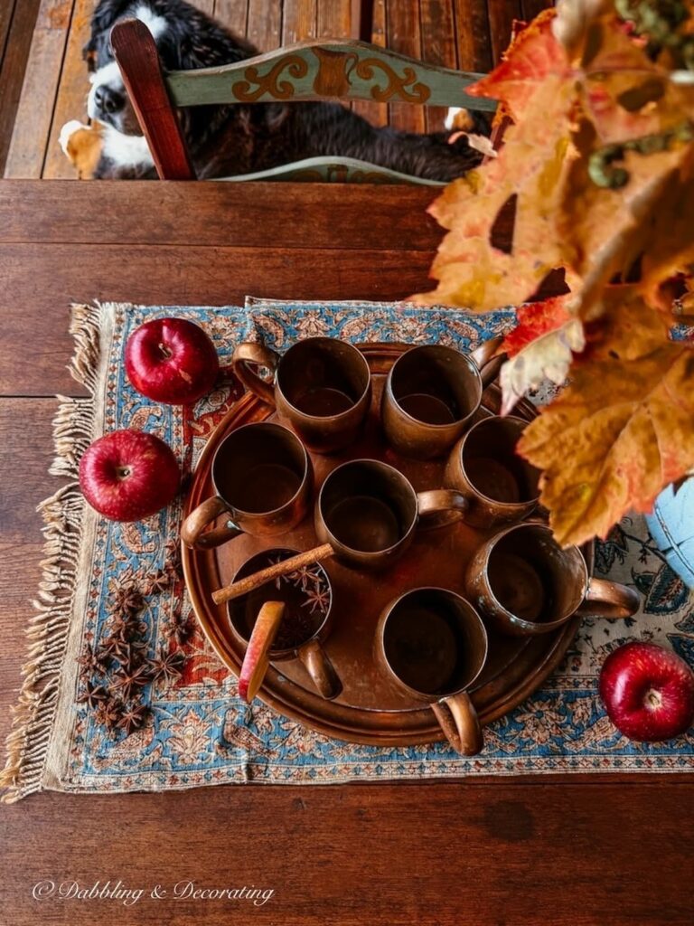 Fall porch table with a set of vintage copper mugs on a tray with apples, antique Iranian textile, foraged centerpiece and a Bernese Mountain Dog looking on.