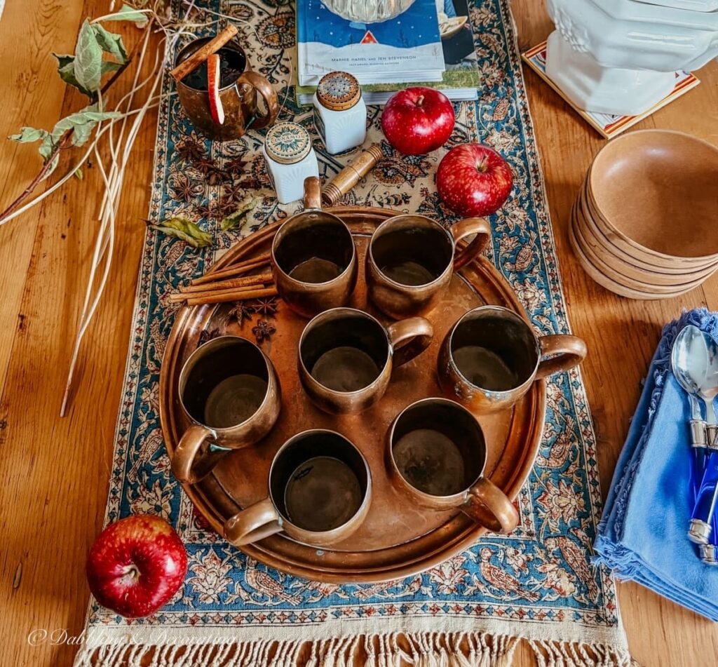 Fall table setting with vintage copper mugs, tray, apples, antique textile, wooden soup bowls, branches, and more for an after apple picking gathering.