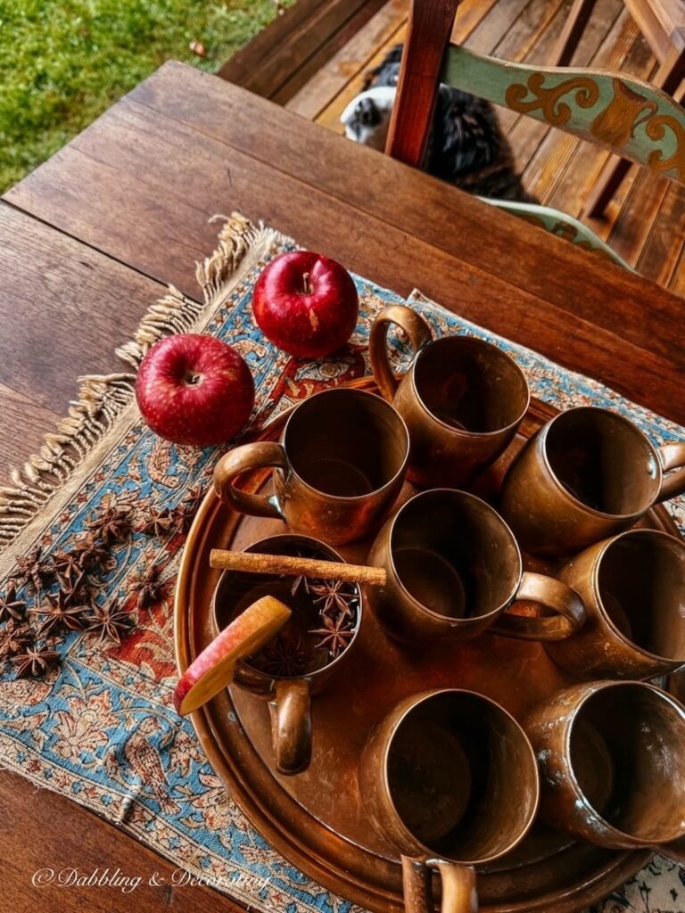 A set of vintage copper mugs on a tray paired with an antique Iranian textile and apples on an outdoor fall porch table with a Bernese Mountain Dog underfoot.