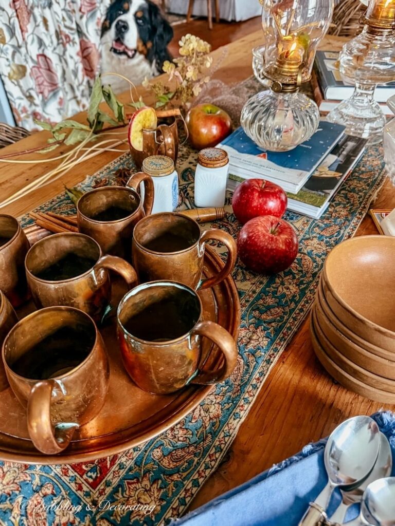 Vintage fall table set for a small gathering with vintage copper, antique blue textile, apples, old spice jars, wooden bowls, books, oil lamps and a Bernese Mountain Dog looking on.