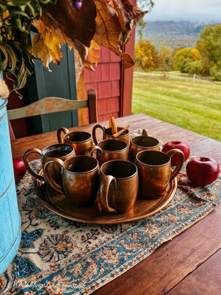 A set of 8 vintage copper mugs on a copper tray on an outdoor fall table with an Iranian antique textile on a Vermont porch during fall foliage with misty mountain views.