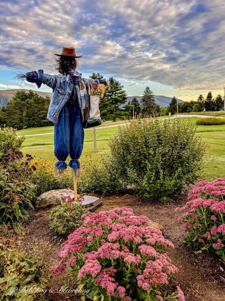 Fall scarecrow in a mountain garden dressed and layered in thrifted blue clothes with a bag full of flowers on the arm.