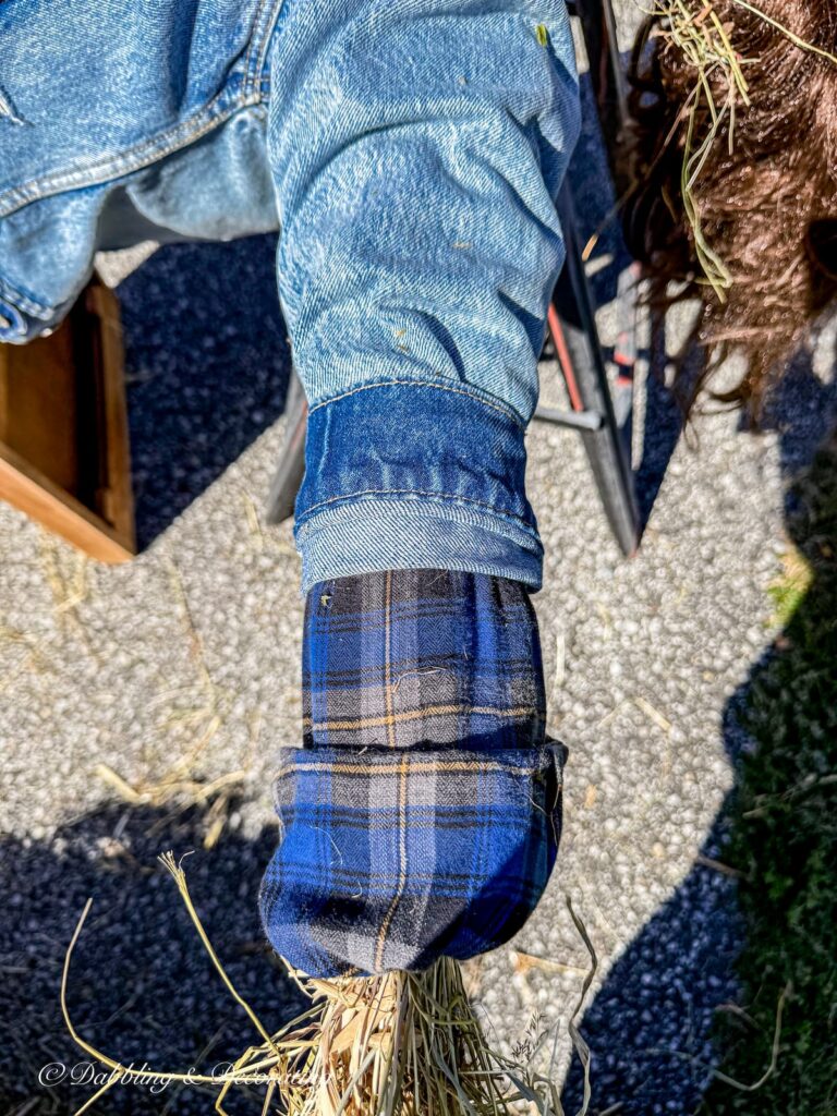 Arm of fall scarecrow layered in blue denim and plaid and stuffed with straw and tied with rubber band to secure it.