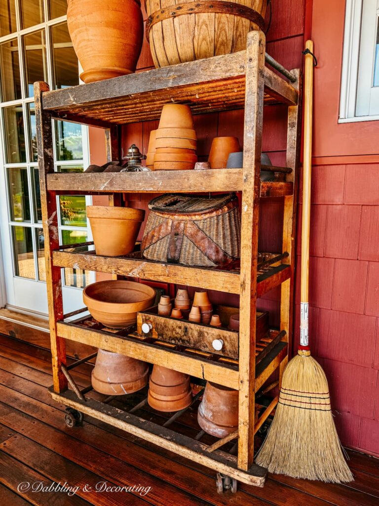 Vintage cobbler rack on colorful fall porch displayed with terracotta pots, fishing basket and one broom leaning up against the baker's rack.