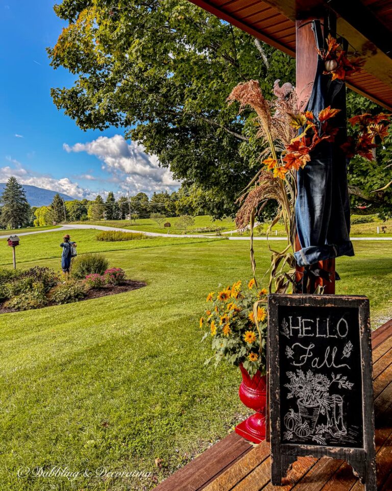 Covered fall porch with Hello Fall chalkboard sign, corn broom, red planters with yellow flowers and mountain views.