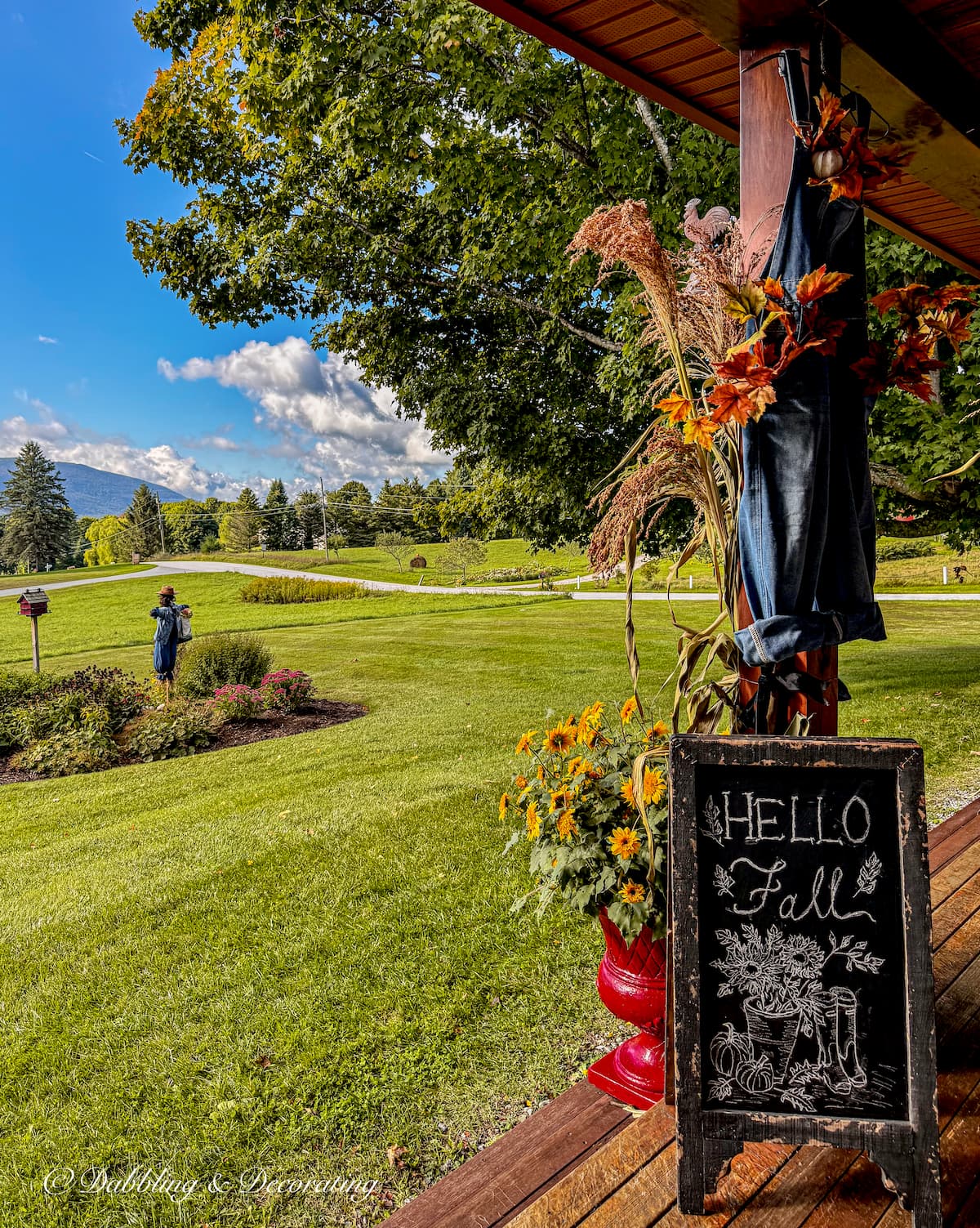 Covered fall porch with Hello Fall chalkboard sign, corn broom, red planters with yellow flowers and mountain views.