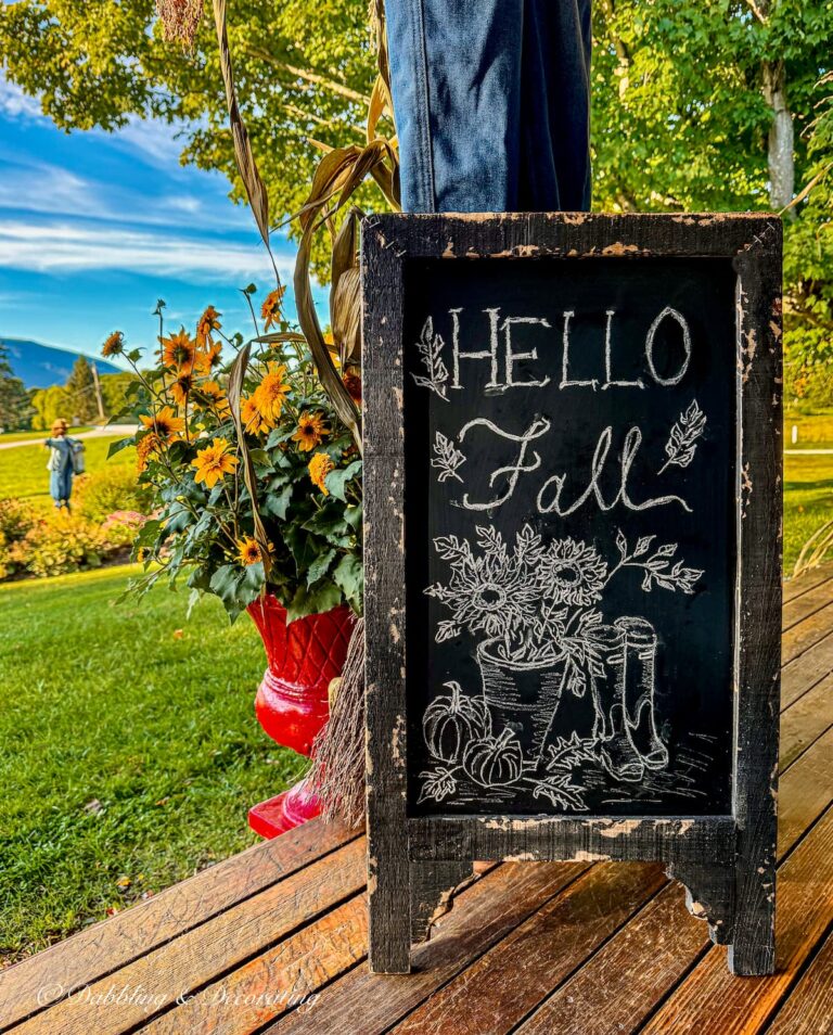 Colorful fall porch with bistro chalkboard sign with Hello Fall artwork, red planter with yellow flowers and changing foliage in the mountains.