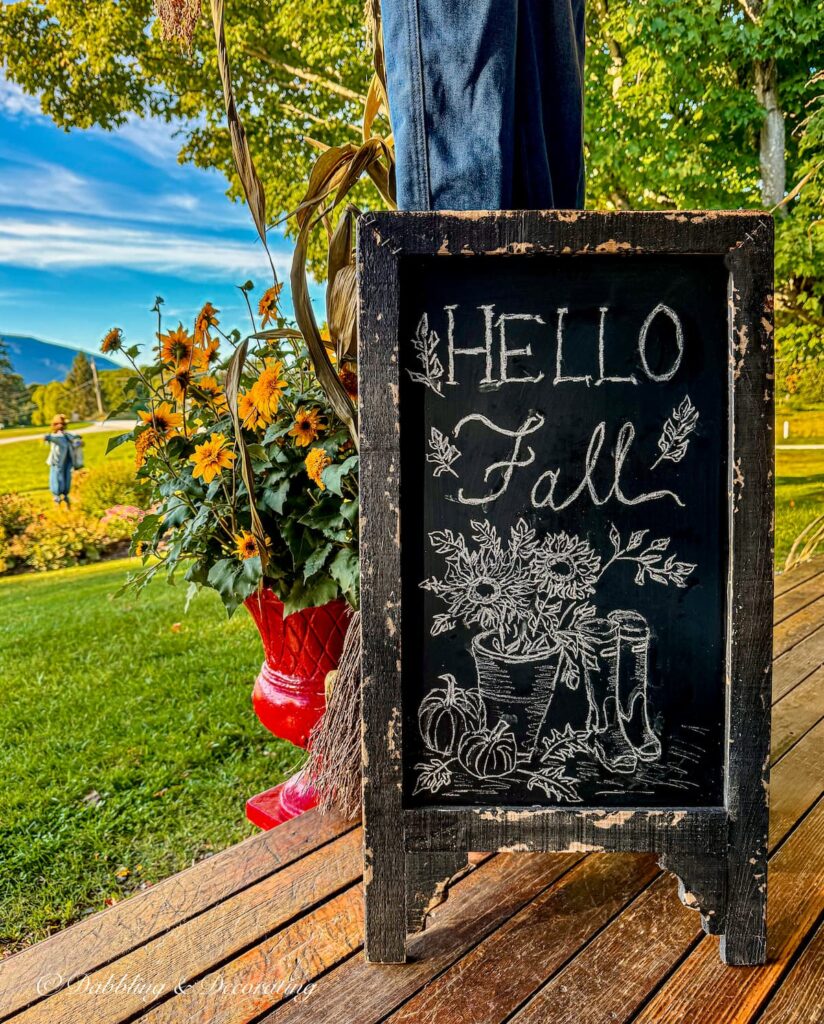 Colorful fall porch with bistro chalkboard sign with Hello Fall artwork, red planter with yellow flowers and changing foliage in the mountains.