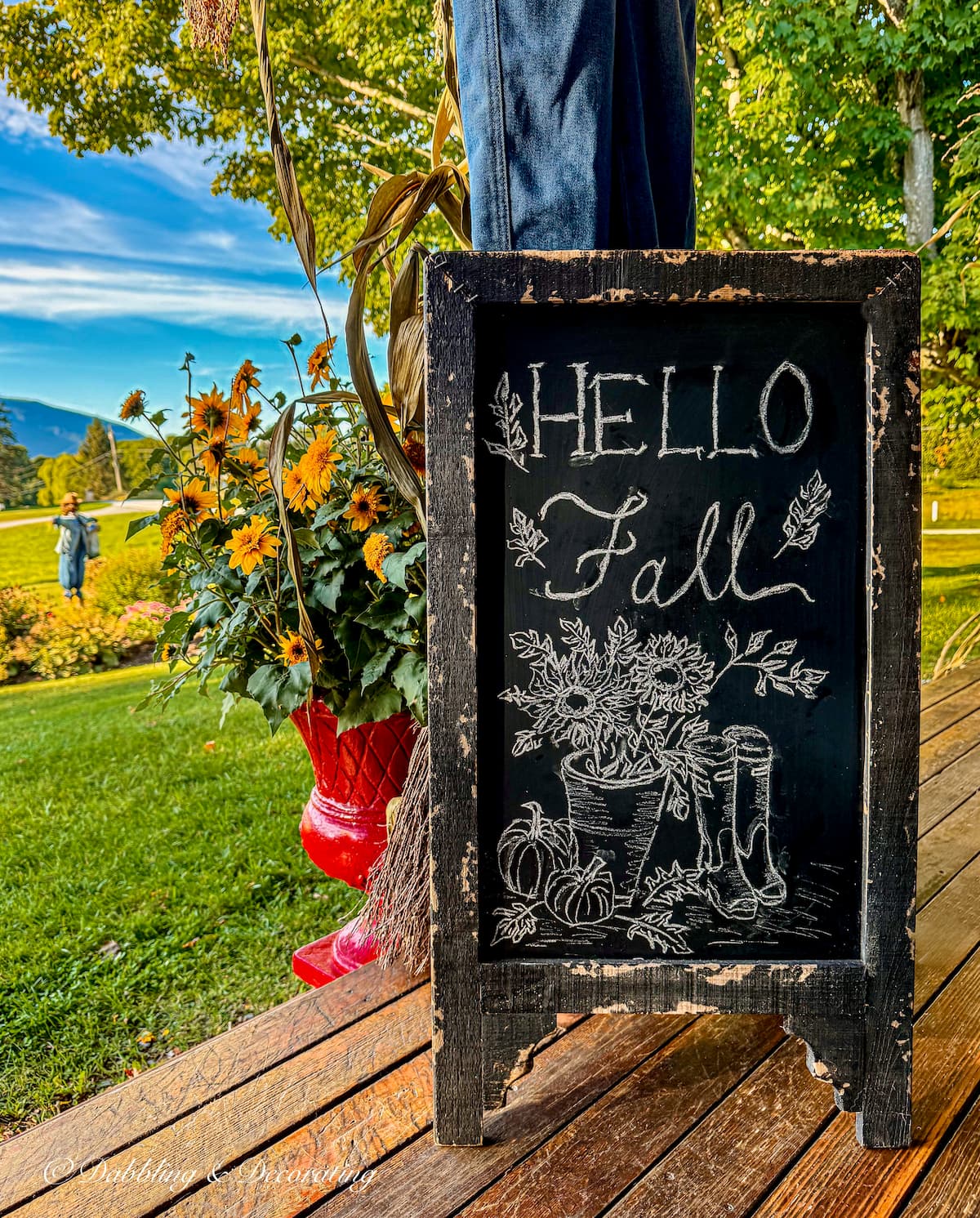 Colorful fall porch with bistro chalkboard sign with Hello Fall artwork, red planter with yellow flowers and changing foliage in the mountains.