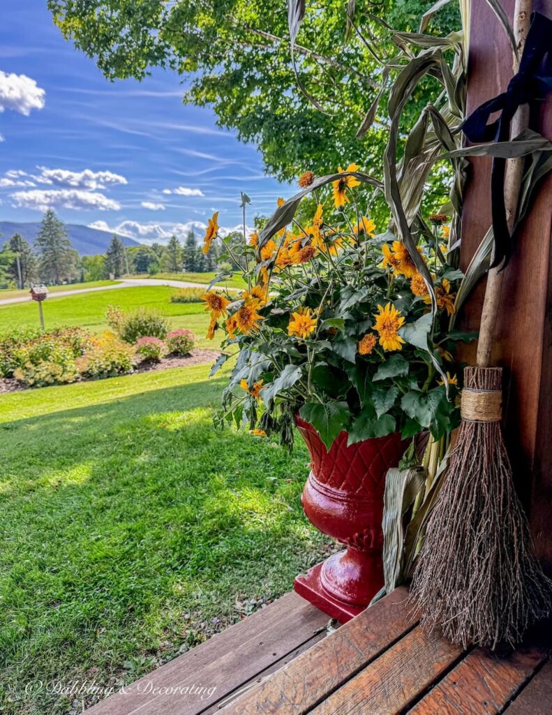 Red planter with whimsical yellow flowers like daisies on colorful fall porch with witches broom attached to beam with black velvet ribbon and mountain views.