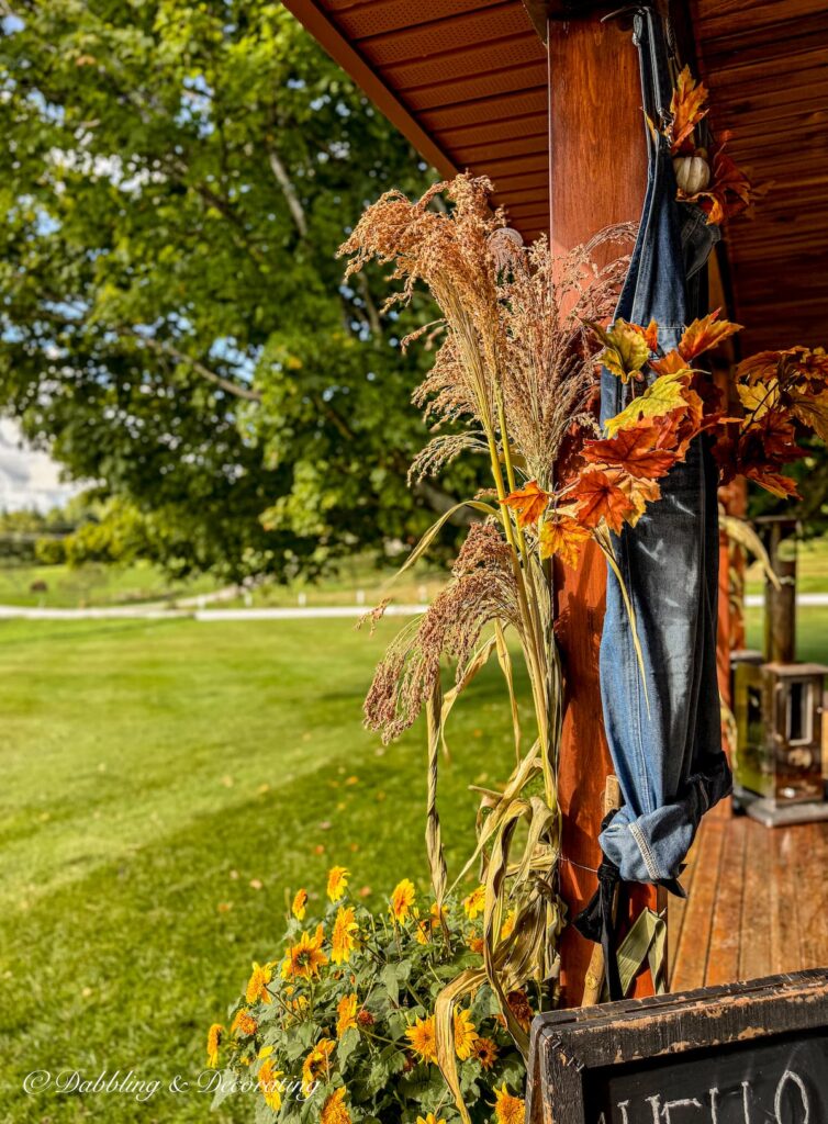 Colorful fall porch with corn broom and overalls tied to a porch beam, yellow flowers in red planters set amongst the Vermont mountains.