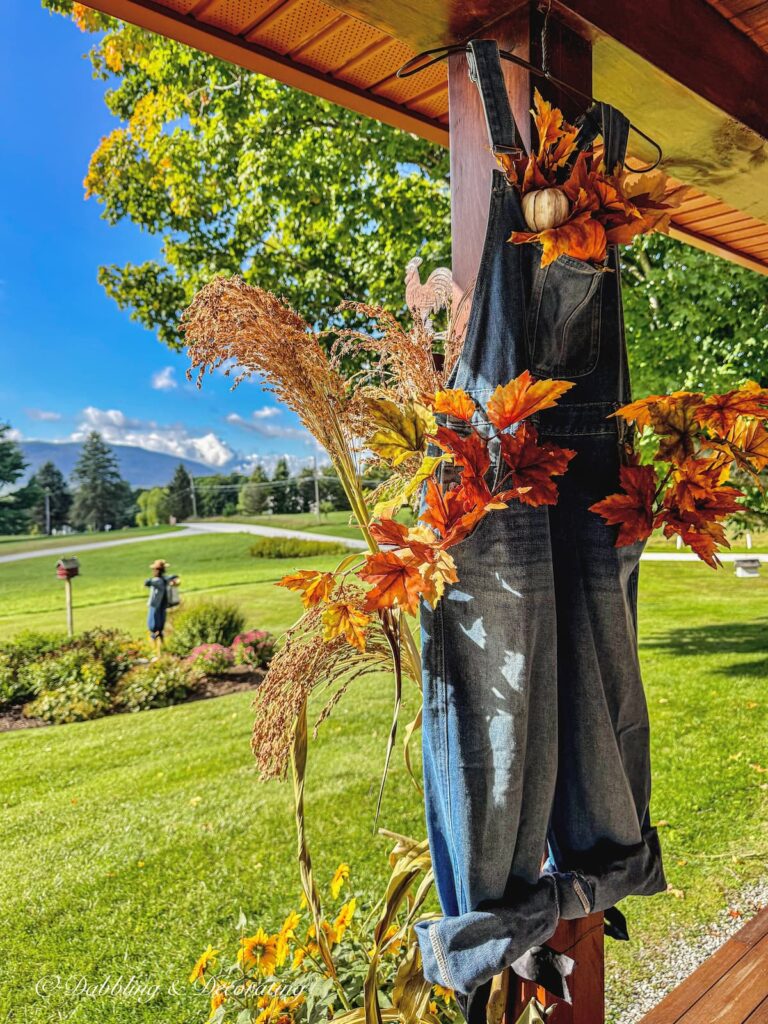 A pair of denim overalls hanging on a colorful fall porch beach stuffed with orange leaves and one orange pumpkin paired with corn broom and yellow flowers on step with mountain views in the background.
