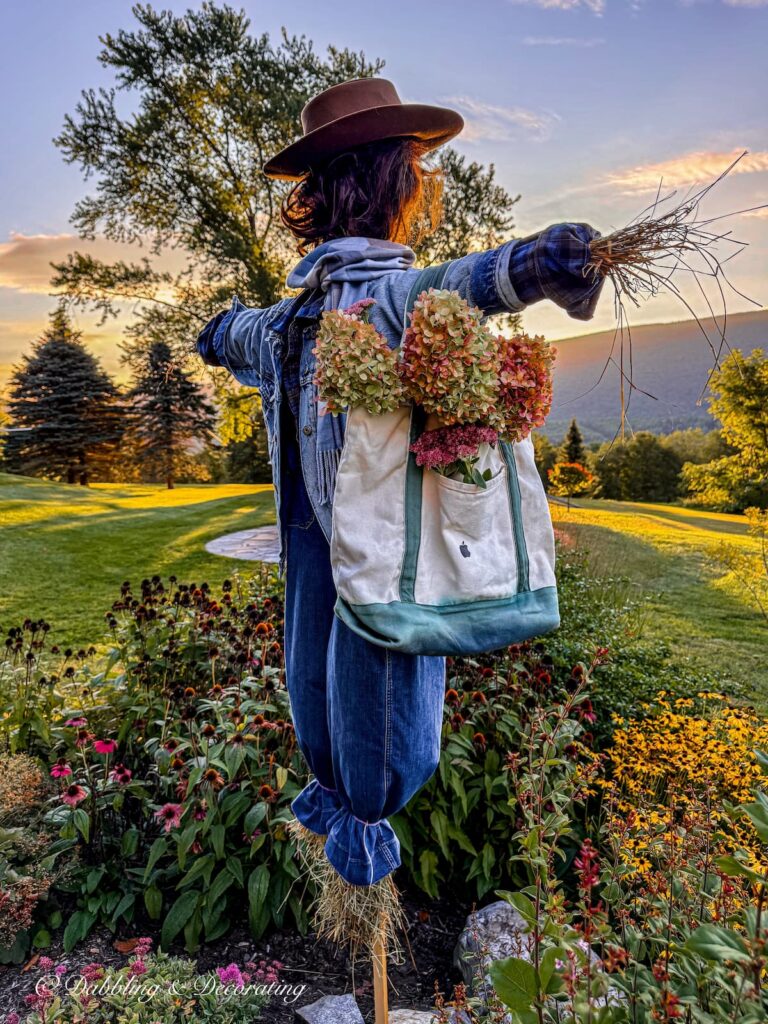 Fall scarecrow layered in denim and thrifted blue with a bag full of fall blooms in a mountain garden with views at sunrise.