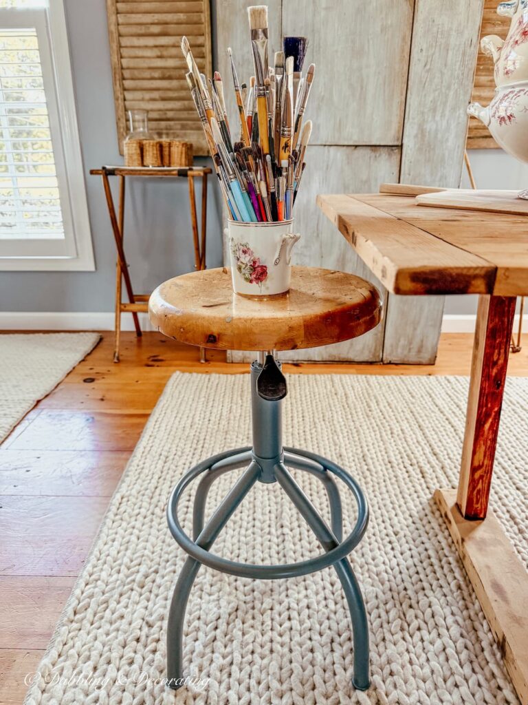 Antique drafting stool restoration with blue-gray paint finished with ceramic jar filled with paintbrushes in kitchen scullery.