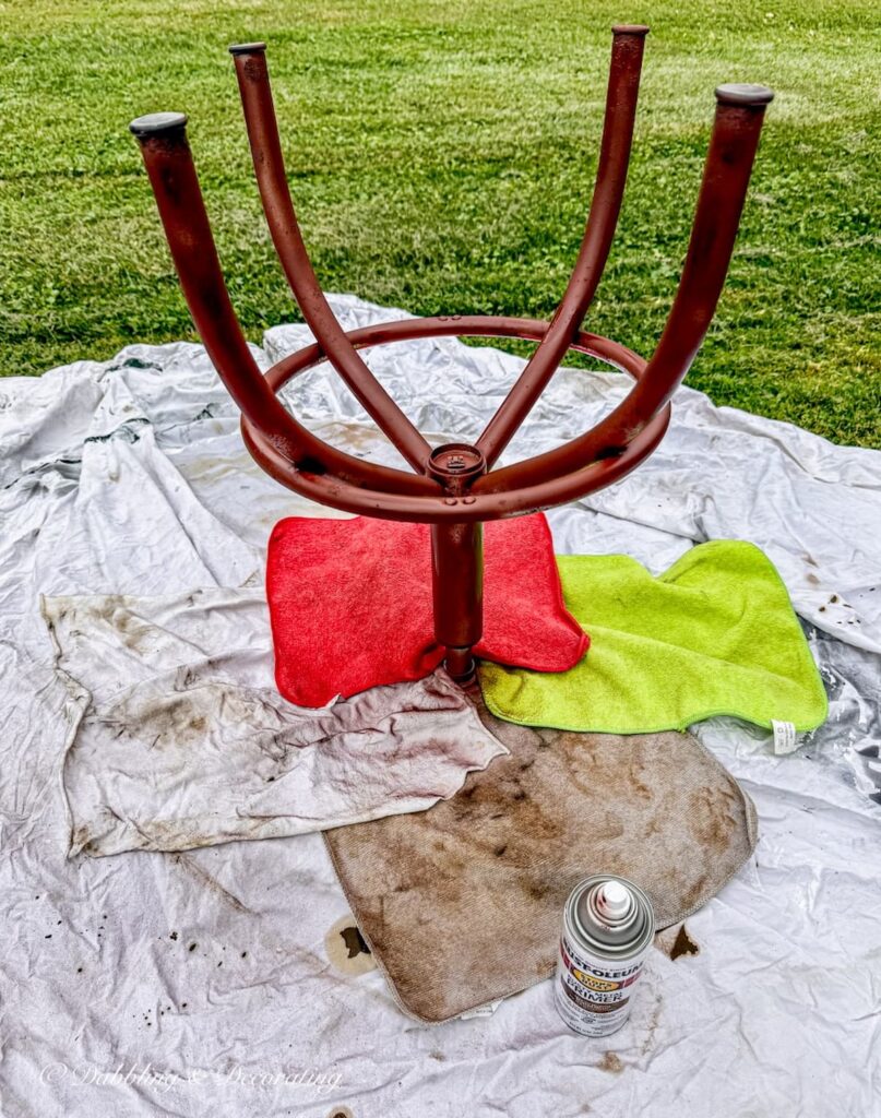 Antique drafting stool upside down covered in rags and legs primed with Rust-Oleum Rusty Metal Primerin brown drying.