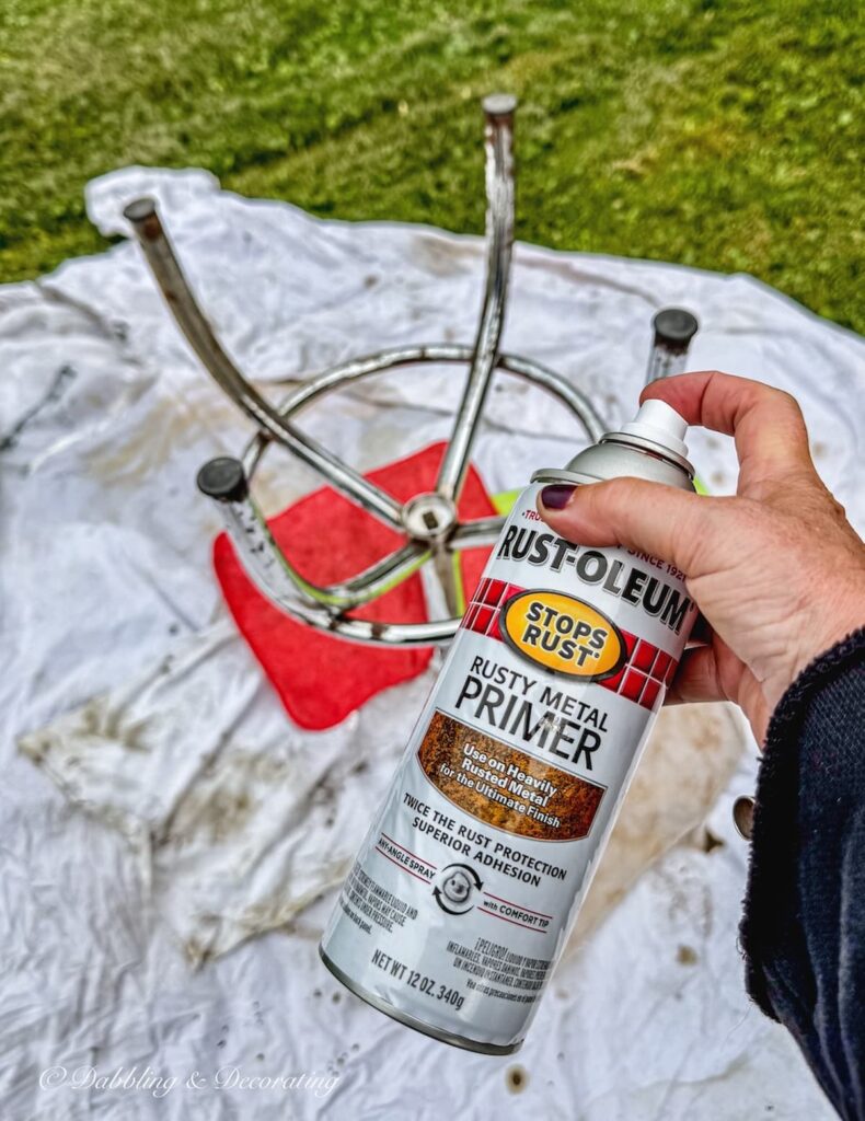 Upside down drafting stool with metal legs ready to prime and wooden stool top covered in rags and Rust-Oleum Rusty Metal Primer in hand.