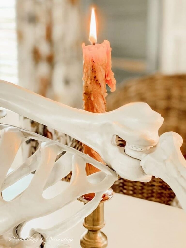 Vintage brass candlestick with drippy orange candle lit next to skeleton on dining room table close up.