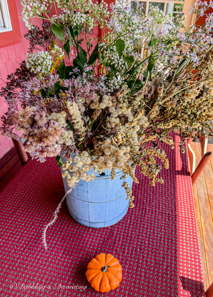 Fall foraged arrangement in vintage blue bucket on colorful fall porch table with small pumpkin.