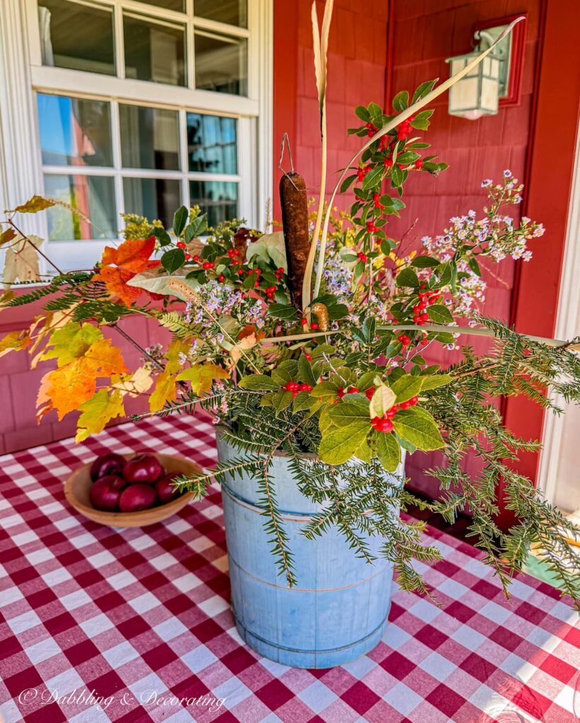 Foraged fall arrangement with colorful leaves, wildflowers, and evergreen in vintage blue bucket on red and white gingham tablecloth on covered porch table.