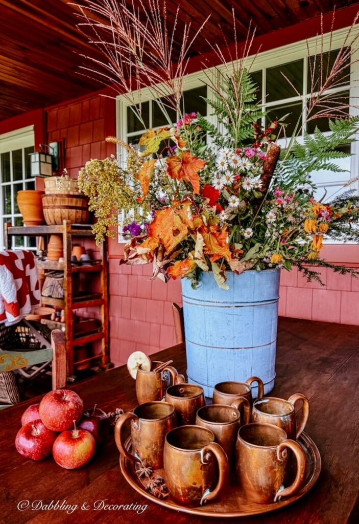 Colorful foraged fall arrangement in a vintage blue ice cream bucket on covered porch table with a tray of vintage copper mugs for fall styling.