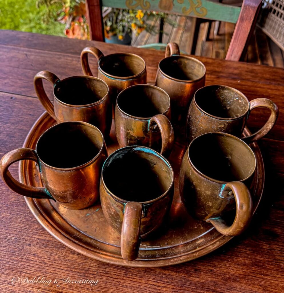 A set of 8 vintage copper mugs on a copper tray on an outdoor covered porch wooden table.