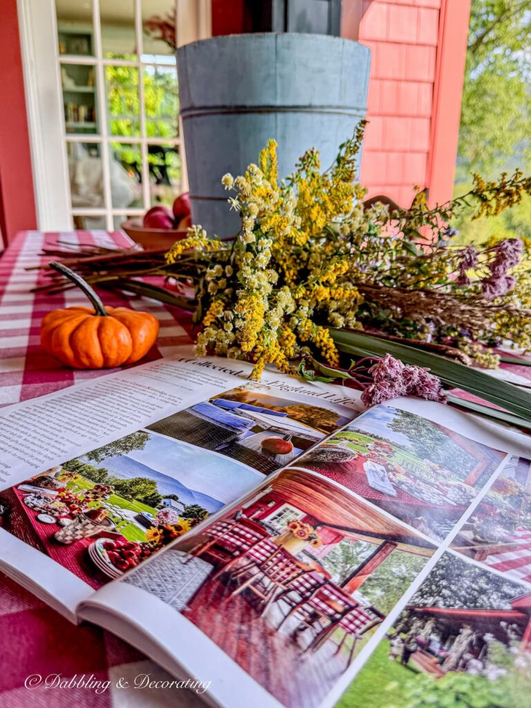 Vintage blue wooden bucket with open Cottages & Bungalows Magazine with fall foraged arrangement and pumpkin on porch table.