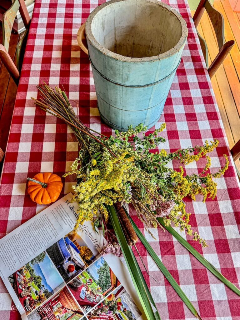 Red and white gingham tablecloth with a vintage blue ice cream bucket, foraged fall flowers, open magazine and pumpkin on porch table.