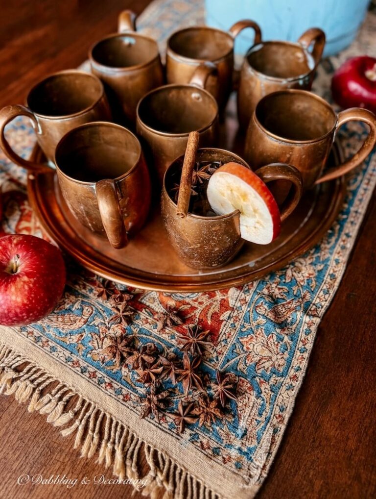 Antique Iranian textile on wooden fall table with vintage copper mugs on tray, slices of apple, and spices for a fall vignette on the porch.