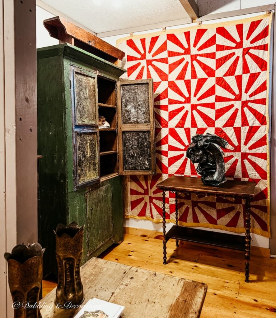 Colorful red antique quilt as wall art next to a primitive antique green hutch in High-End Antique Booths inside Taconic Antiques Fair.
