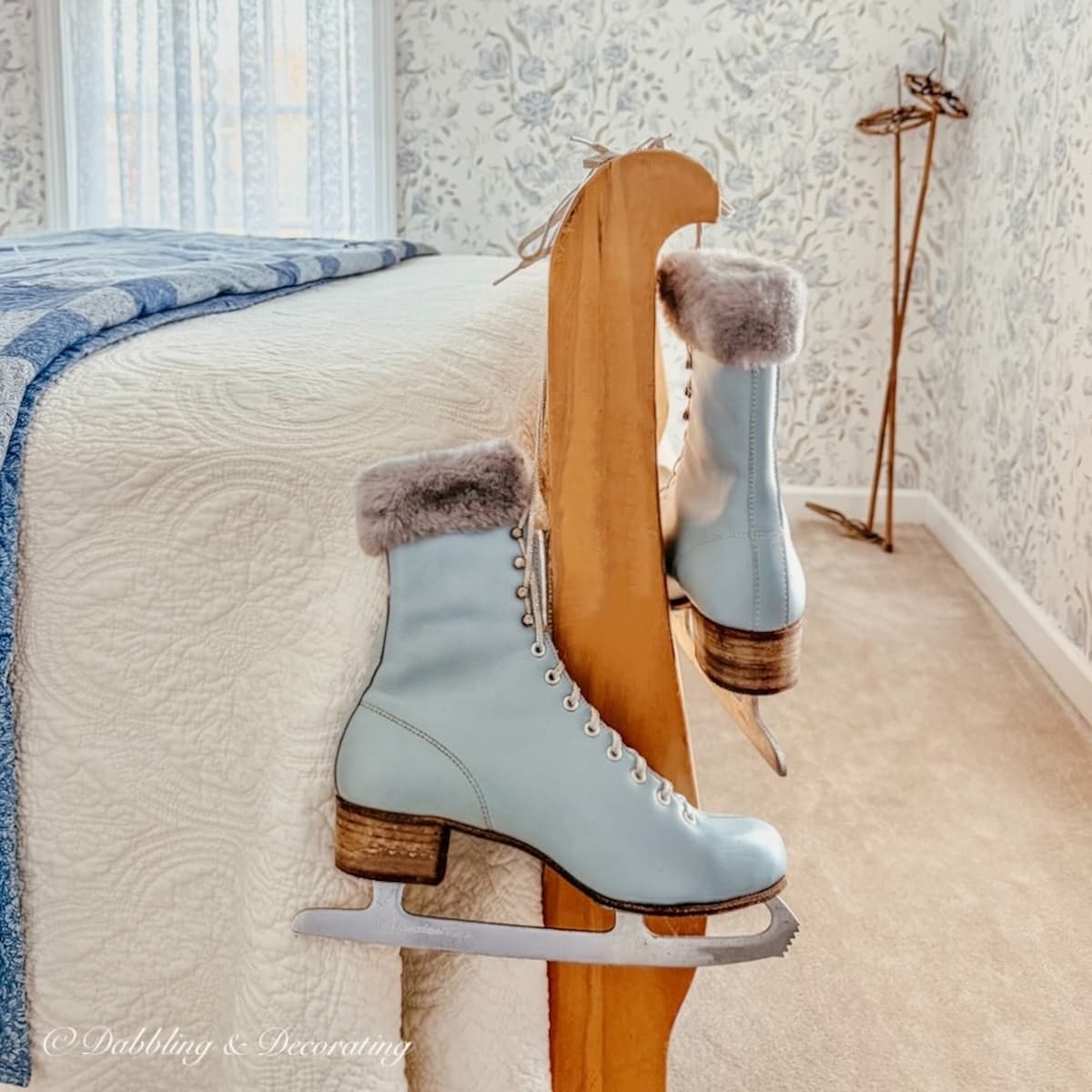 Vintage blue ice skates with ankle fur hanging from a vintage pine sleigh bed in bedroom with wallpaper in blue and cream color scheme.