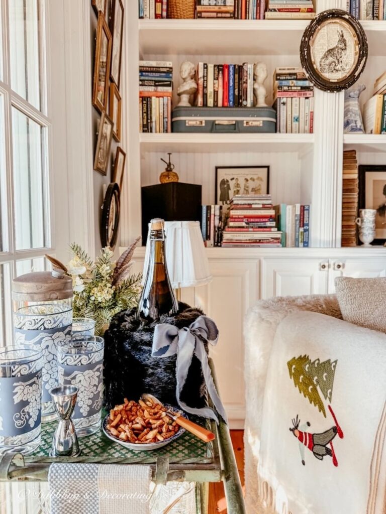 Vintage black hand muff as a wine cover on a vintage bar cart in front of eclectic styled built-in bookshelves in corner.