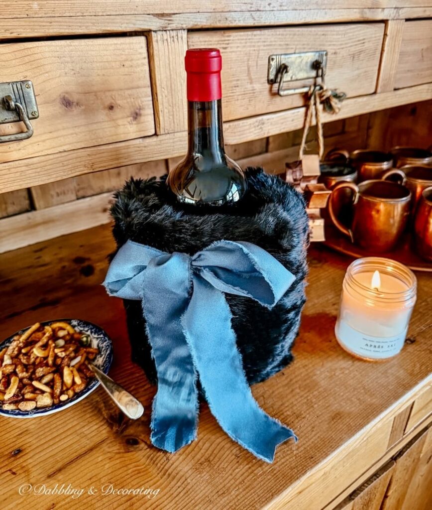 Thrifted black hand muff being used as a wine cover with a blue bow on an antique hutch ledge with lit candle and a bowl of snacks.