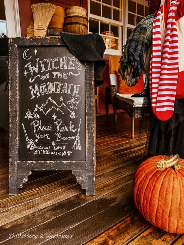 Black bistro chalkboard with Witches on the Mountains Drawing and welcome sign next to a mannequin witch and pumpkin on front porch for Halloween.