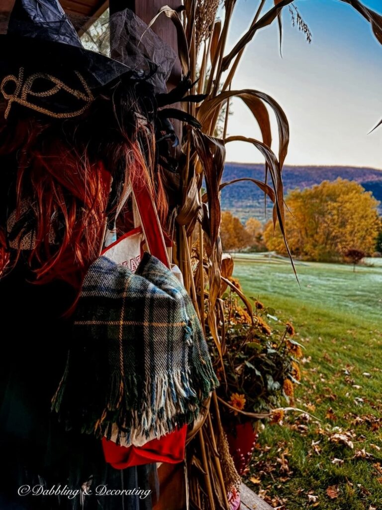 Mannequin dressed as a witch with a red wig, witches hat, mask, and overnight canvas bag with blanket on front porch with corn brooms and fall foliage with mountain backdrops.